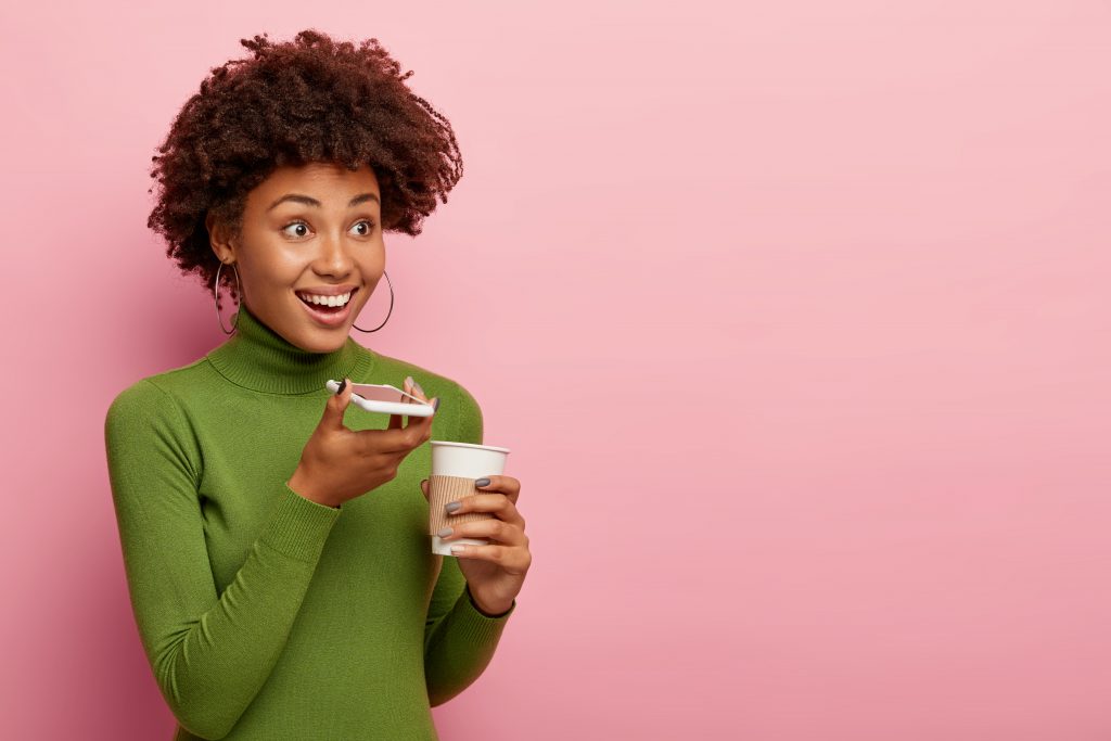 cheerful woman uses voice command recorder holds smartphone device near mouth enjoys drinking aromatic beverage wears green turtleneck